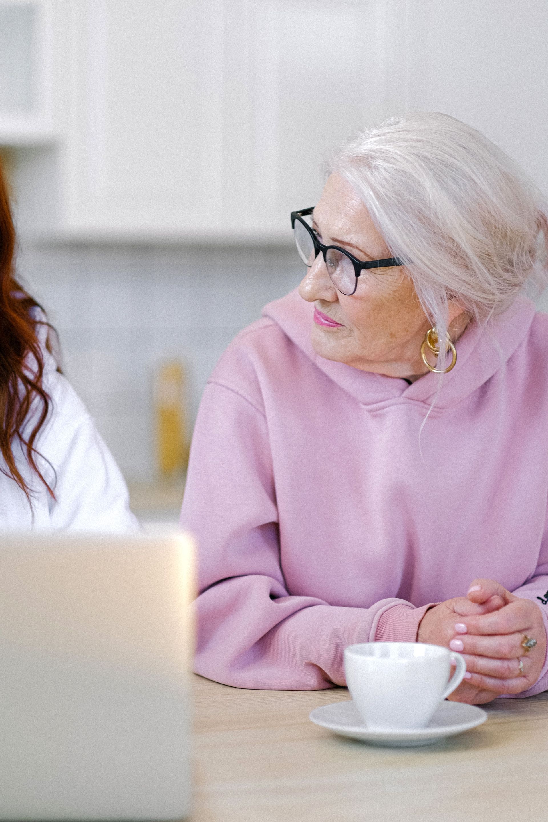 Thoughtful woman with gray hair in eyeglasses and hoodie sitting at table with cup pf tea while spending time with friend in light apartment