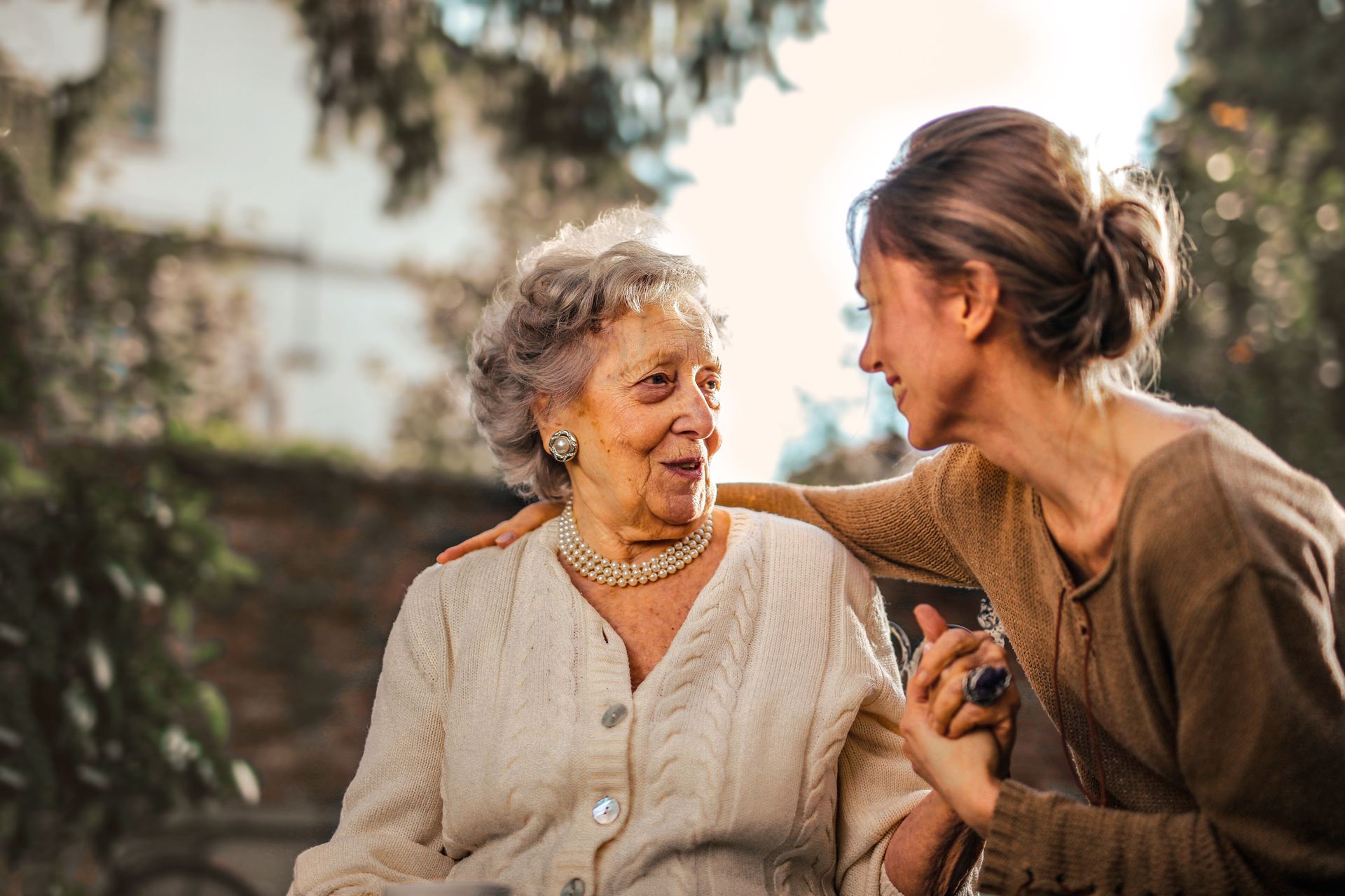 Low angle of cheerful aged lady and adult woman in casual clothes holding hands and smiling at each other while having meeting on yard against blurred green trees and facade of country house in suburb