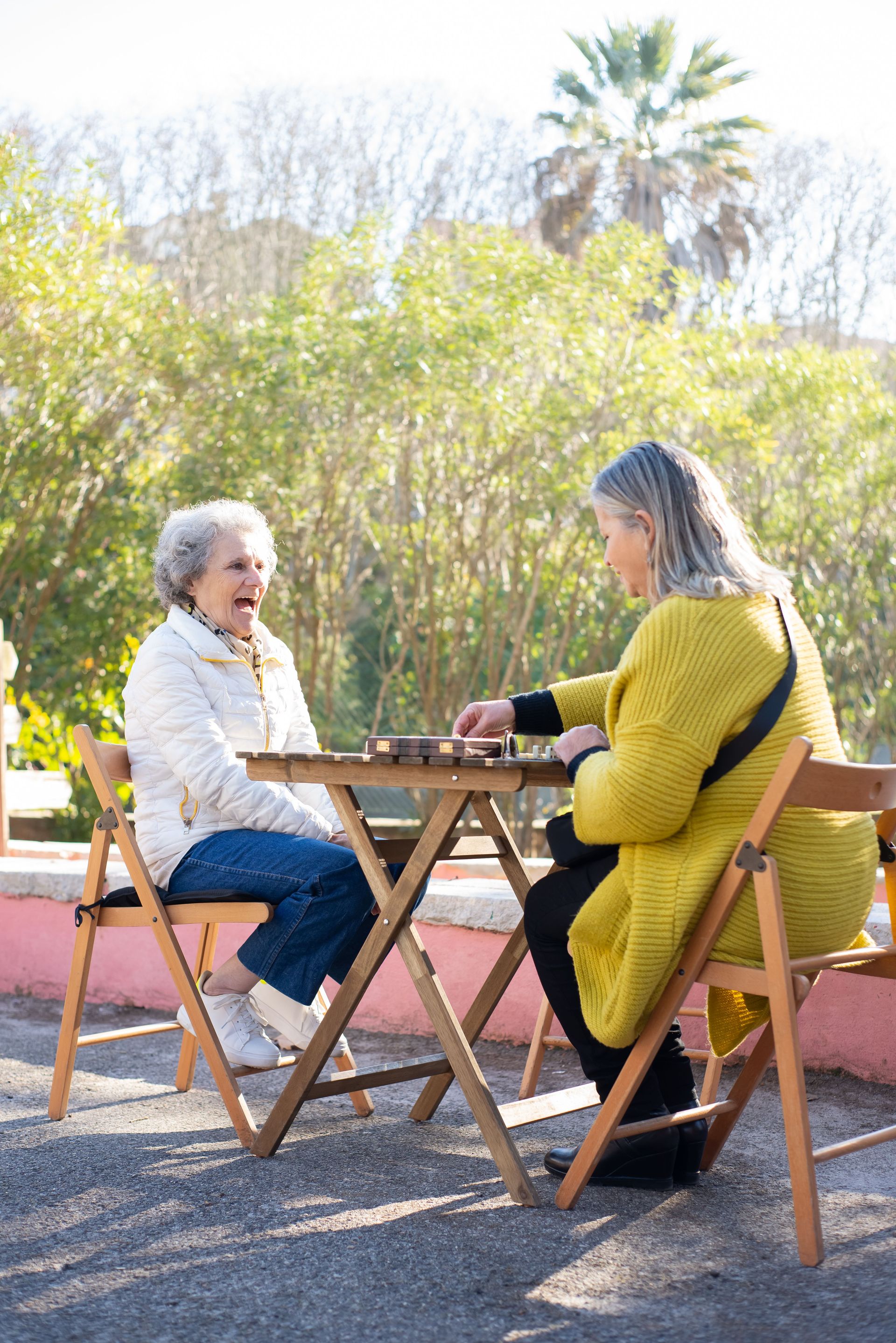 Two senior women laughing and playing dominoes outdoors on a sunny day.